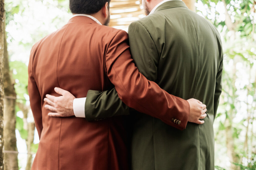 Two grooms in rust and olive suits embracing at Las Caletas wedding ceremony, Puerto Vallarta