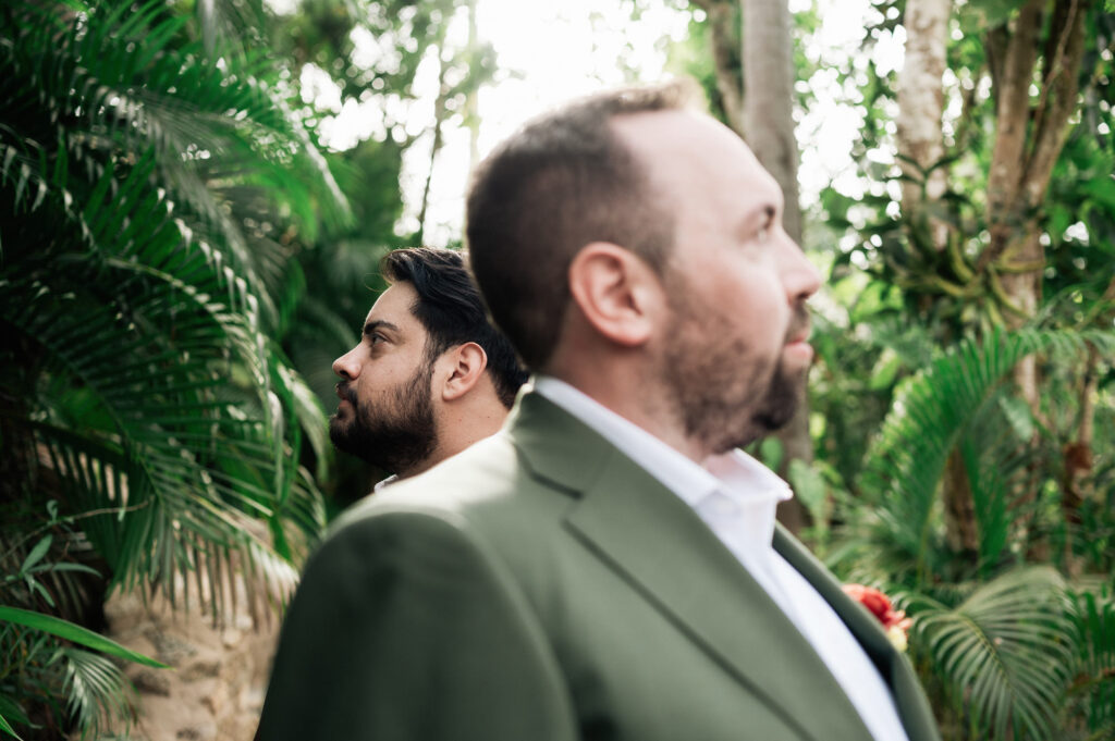 Two grooms in sage green suits share a quiet moment among tropical palms at Las Caletas, Puerto Vallarta wedding