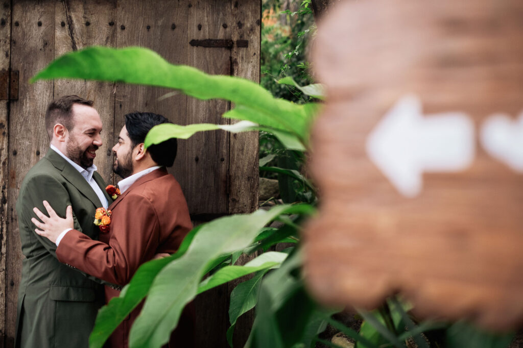 Two grooms laughing together at Las Caletas Puerto Vallarta wedding, photographed through tropical leaves