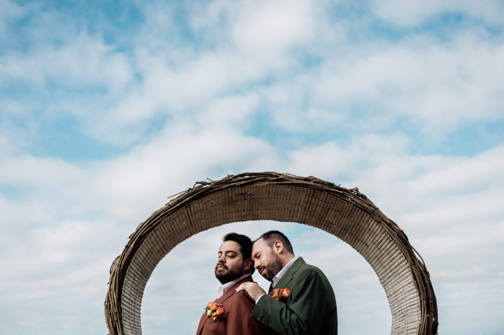 Two grooms in burgundy and green suits share a tender moment under a circular arch at Las Caletas, Puerto Vallarta wedding