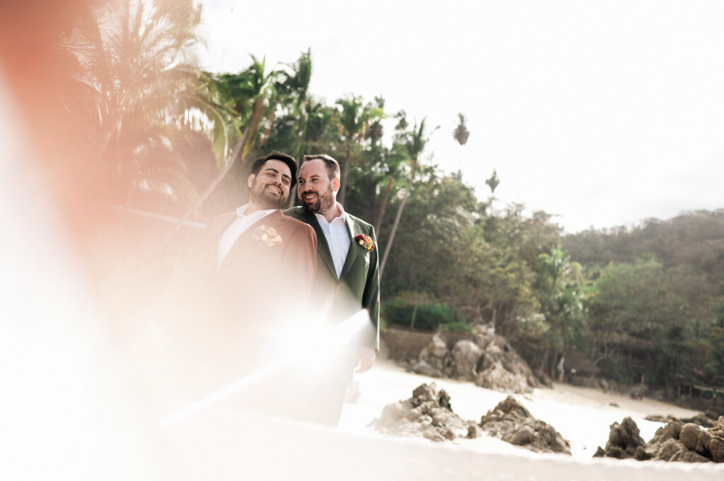 Two grooms in suits with boutonnieres embrace on the beach at Las Caletas, Puerto Vallarta wedding