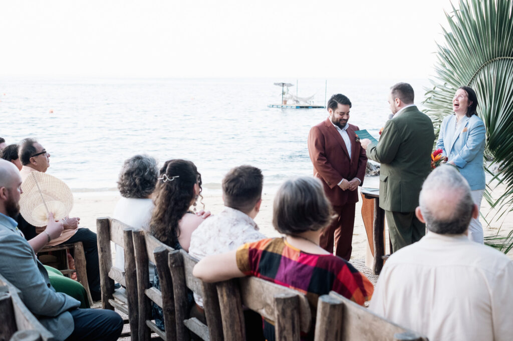 Beach wedding ceremony at Las Caletas Puerto Vallarta with two grooms and laughing officiant