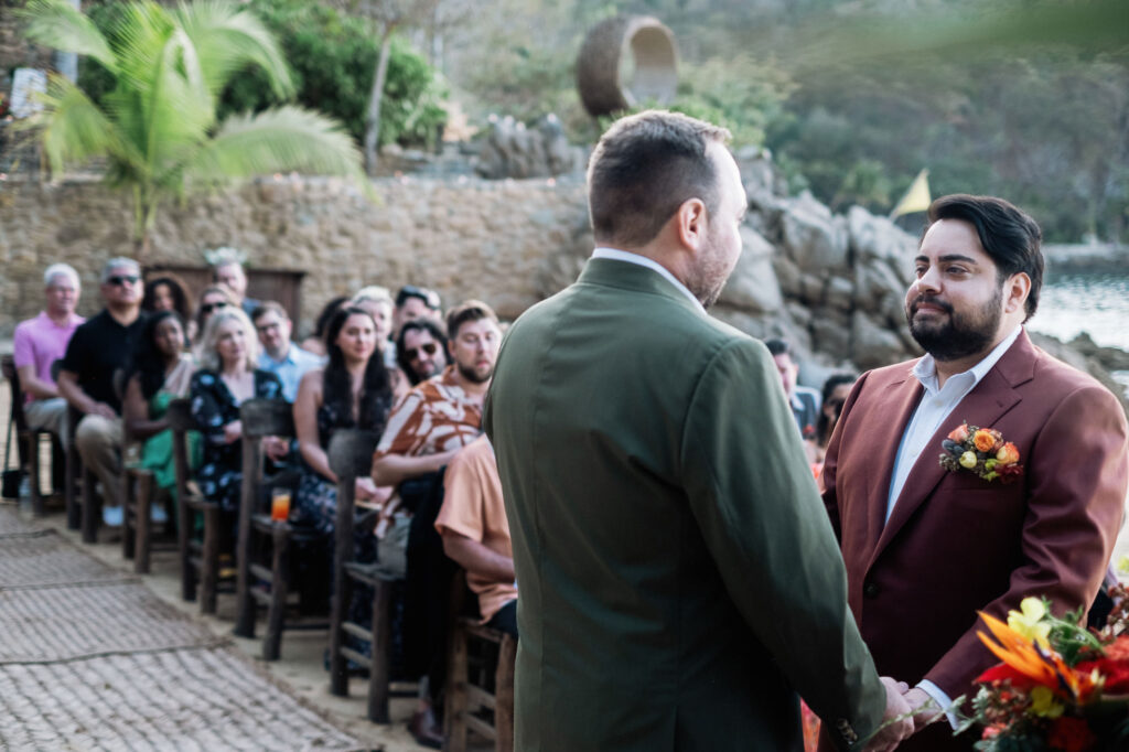Two grooms face each other during outdoor wedding ceremony at Las Caletas Puerto Vallarta with guests seated behind