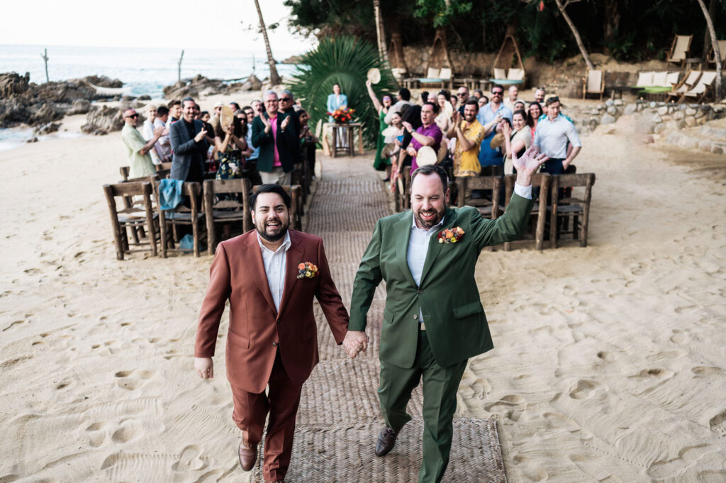 Two grooms walk down beach aisle holding hands at Las Caletas wedding, Puerto Vallarta, guests cheering behind them