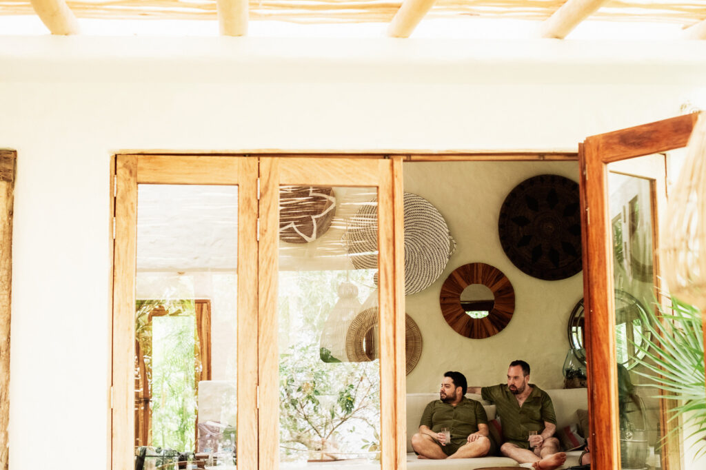 Two grooms relaxing together at Las Caletas Puerto Vallarta wedding, holding drinks in a boho villa