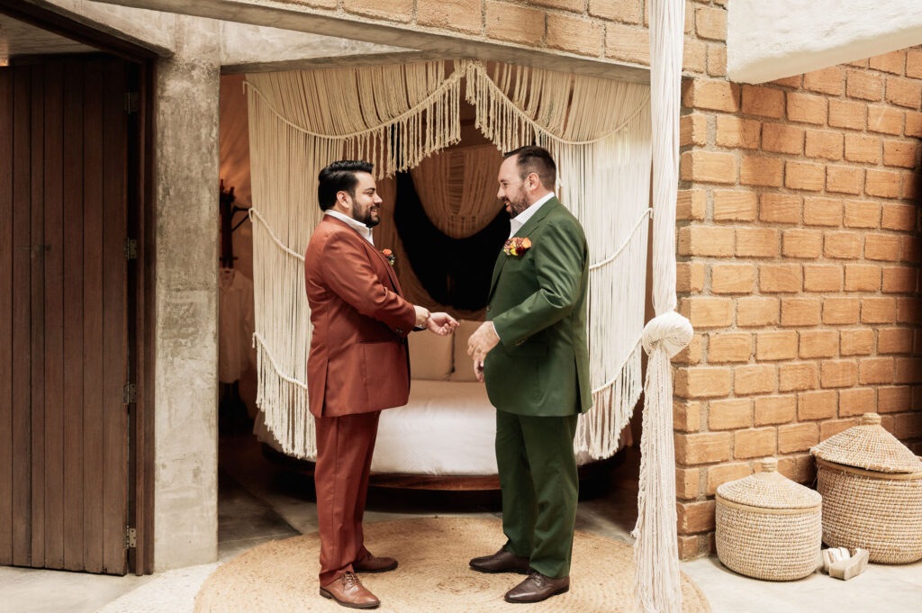 Two grooms in terracotta and green suits hold hands laughing at Las Caletas wedding, Puerto Vallarta