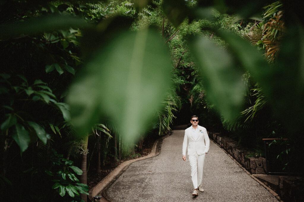 Groom in white suit walking through lush tropical path at One&Only Mandarina wedding, Mexico