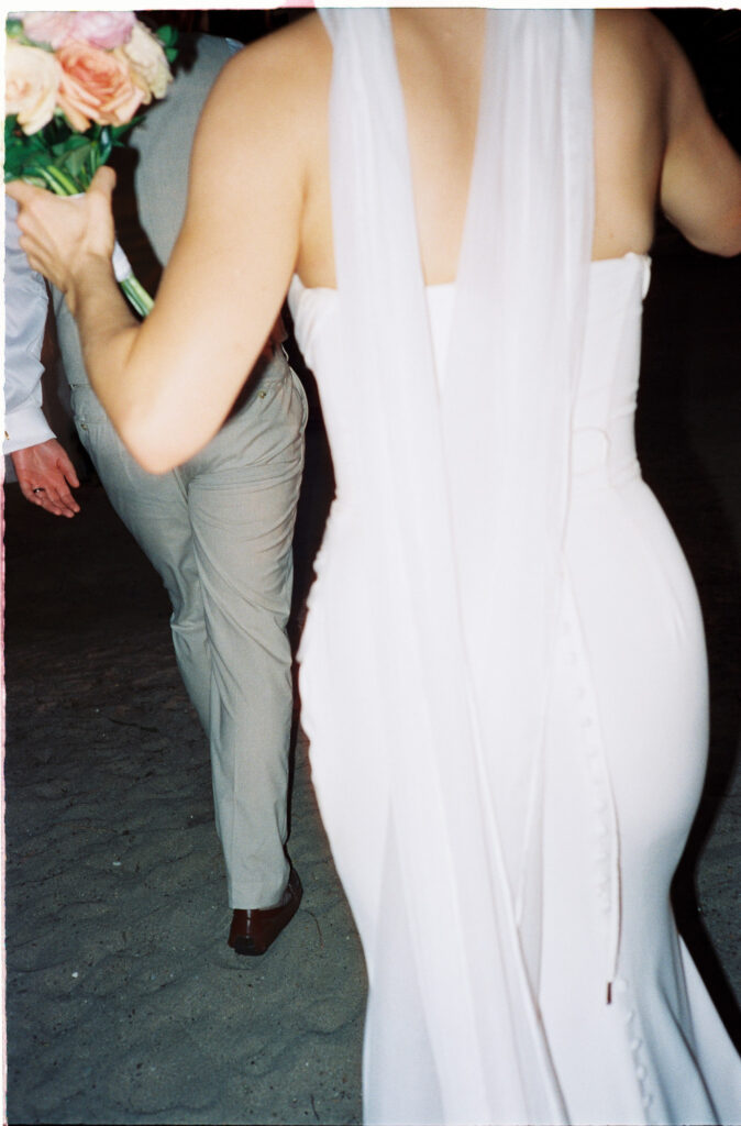 Bride in backless white gown holding bouquet walks with groom on sandy beach at Riu Vallarta wedding reception