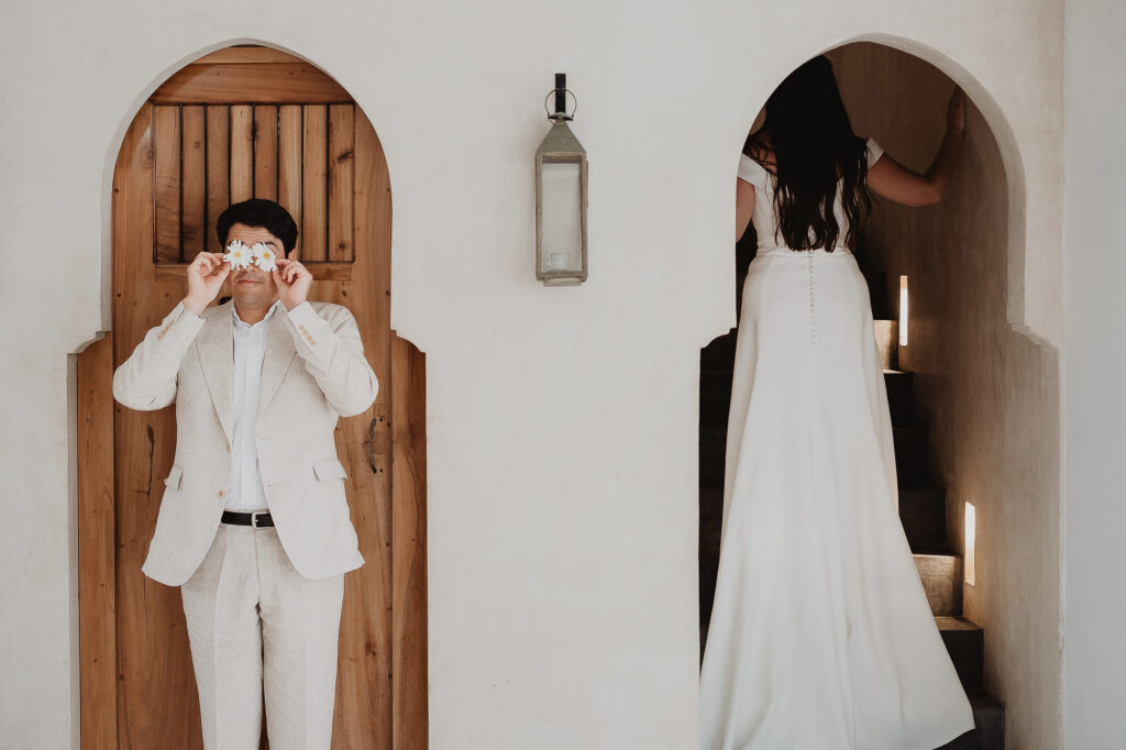 Groom holds daisies over his eyes while bride walks upstairs at Ambre Epieces Tulum wedding