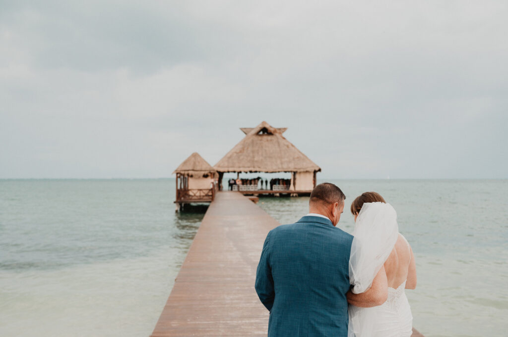 Bride and groom walking the pier toward overwater palapa at Dreams Vista Cancun wedding ceremony