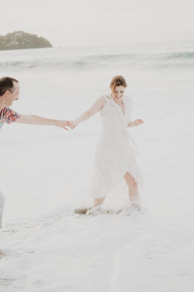 Couple laughing and holding hands in ocean waves during engagement session in Sayulita, Mexico