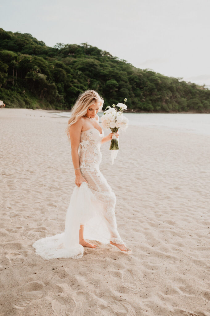 Bride in lace gown holding white bouquet barefoot on beach at One&Only Mandarina elopement