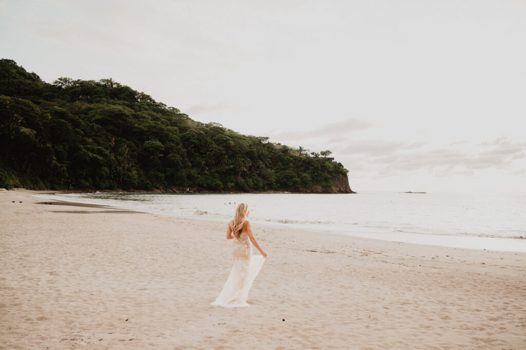 Bride in lace gown walks along beach at One&Only Mandarina during elopement, lush jungle cliffs behind her