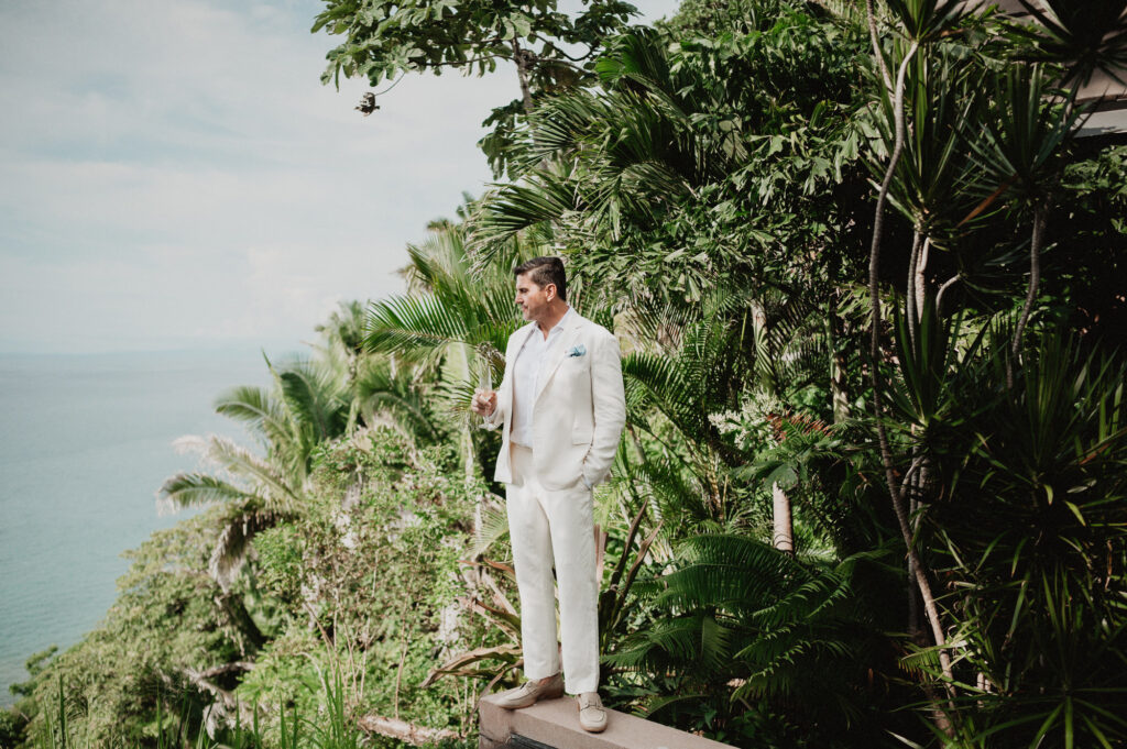 Groom in white linen suit holding champagne at One&Only Mandarina elopement, tropical cliffside ocean view