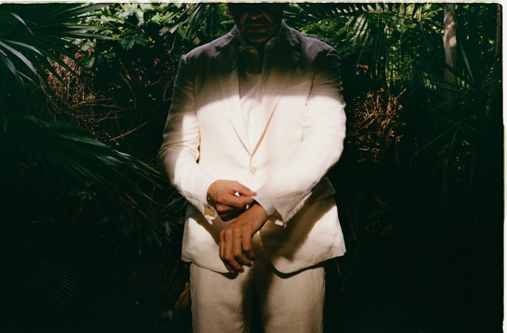 Groom in white linen suit standing among tropical palms at One&Only Mandarina elopement, Mexico