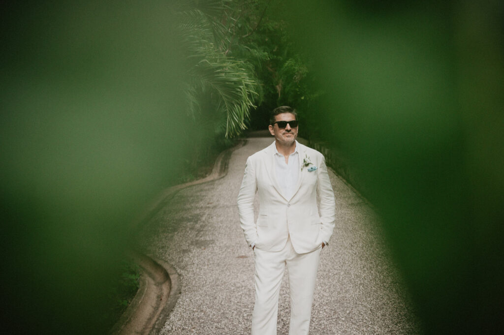 Groom in white linen suit walks garden path at One&Only Mandarina elopement, Mexico