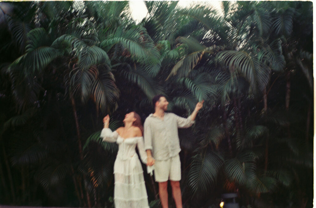 Couple in white holding hands laughing among palm trees, Puerto Vallarta engagement session on 35mm film