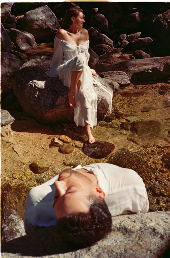 Couple during engagement session on rocky shore in Puerto Vallarta, woman in white dress seated on boulder, man resting below
