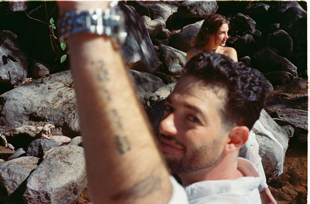 Couple on rocky shore in Puerto Vallarta engagement session, man smiling with tattooed arm raised, analog film