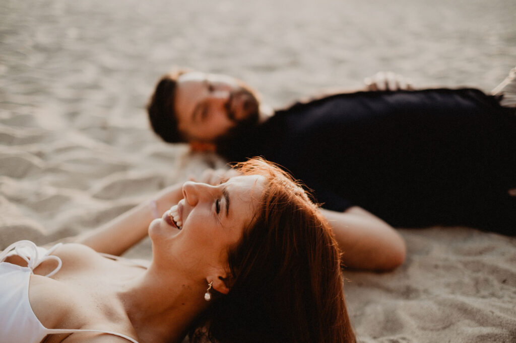 Couple laughing together lying on sandy beach, engagement session in Puerto Vallarta by photographer Alex Obregón