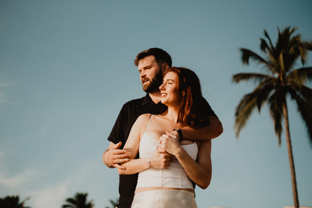Couple embracing at golden hour during engagement session in Puerto Vallarta, palm trees and blue sky