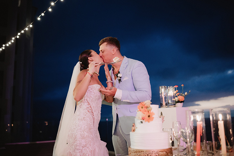 Bride and groom share a kiss while cutting wedding cake at Hilton Riviera Vallarta reception under evening sky