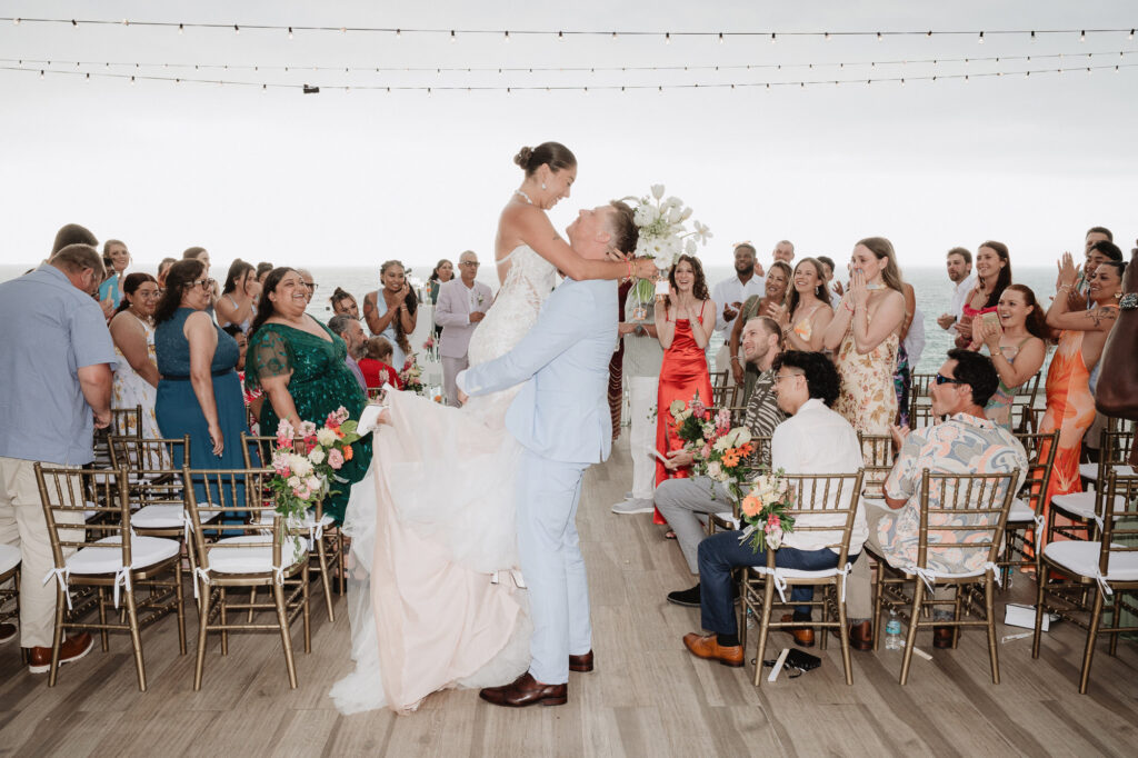 Groom lifts bride during wedding ceremony at Hilton Puerto Vallarta, ocean backdrop, guests cheering