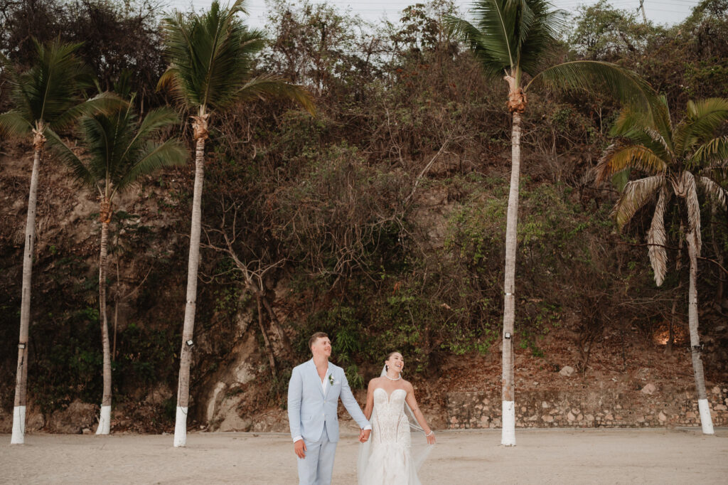 Bride and groom walking hand in hand on beach at Hilton Puerto Vallarta wedding, palm trees behind them