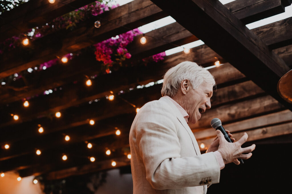 Older man giving wedding speech under pergola with string lights and flowers, San Miguel de Allende reception