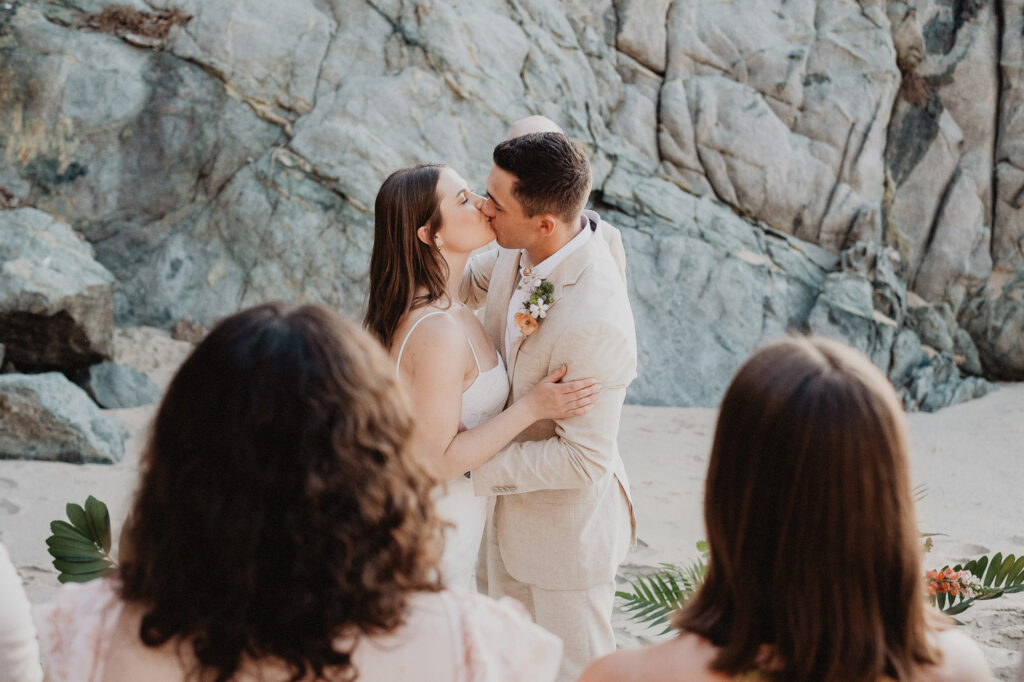 Couple sharing first kiss on sandy beach with rock cliffs, elopement wedding in San Pancho Mexico