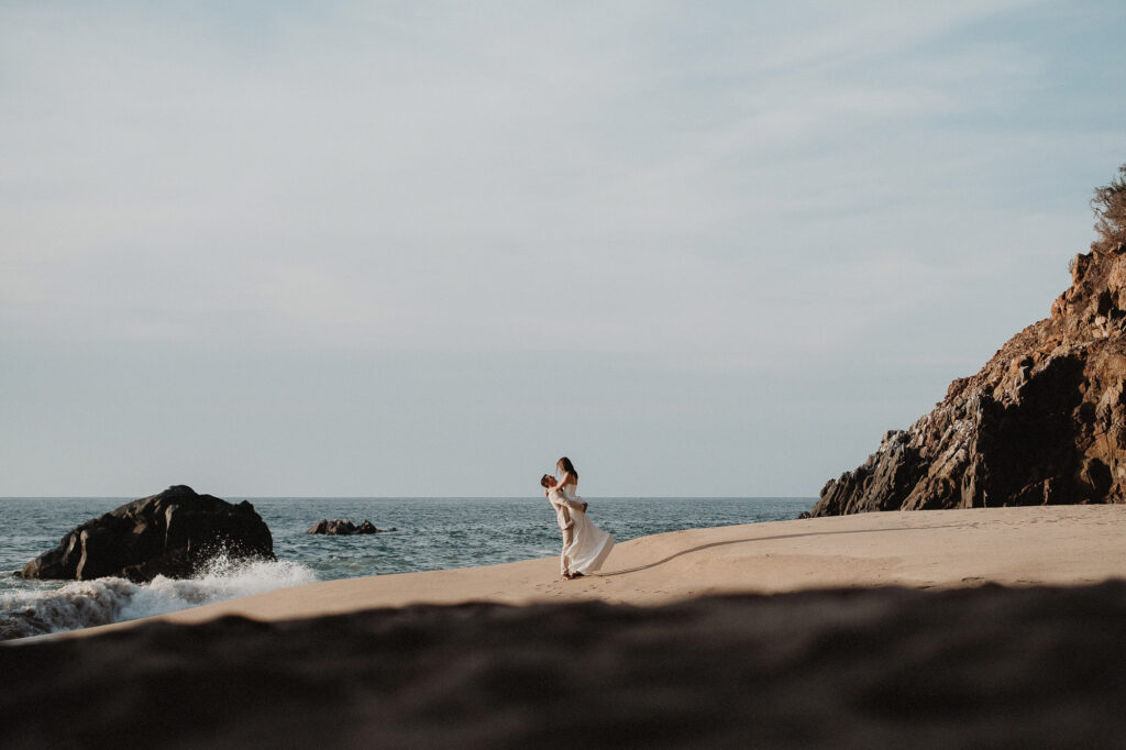 Groom lifting bride on sandy beach at San Pancho elopement, rocky cliffs and ocean waves behind them