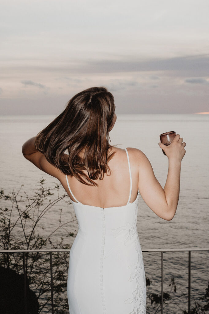 Bride in white gown holds ceramic cup overlooking ocean at sunset, San Pancho elopement photography