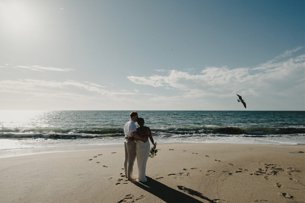 Couple embracing on Sayulita beach at sunset during elopement, bouquet in hand, ocean waves behind them