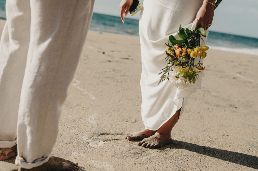 Barefoot bride holding wildflower bouquet beside partner on sandy beach, elopement at Villa Amor Sayulita
