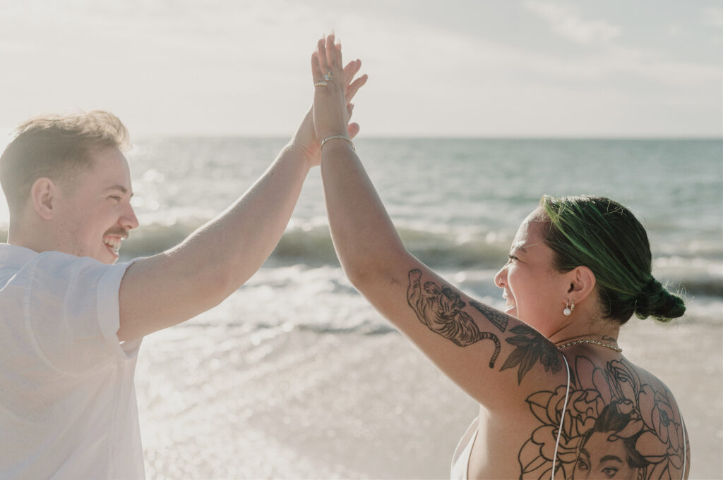 Couple high-fiving and laughing on Sayulita beach during elopement, ocean backdrop, tattooed bride