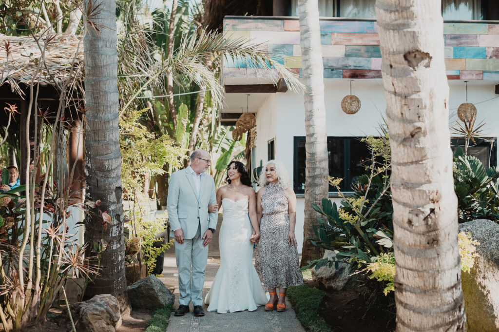 Bride walks with parents through palm-lined path at Frente al Punto, Sayulita wedding ceremony