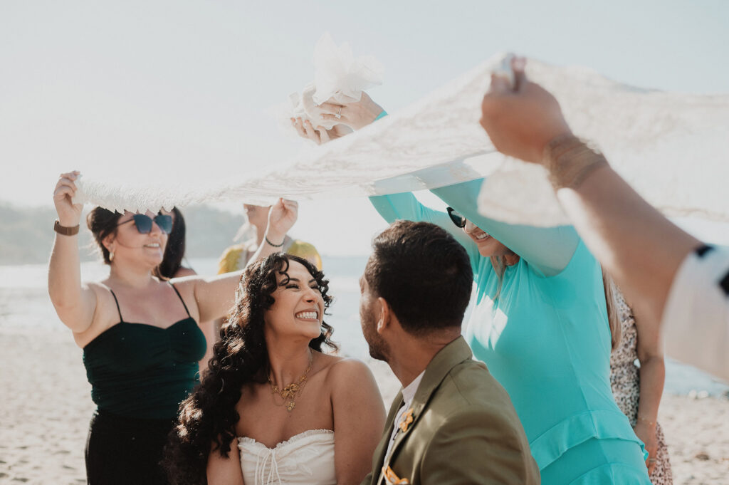 Bride laughing joyfully as guests lift her veil during beach wedding ceremony in Sayulita, Mexico