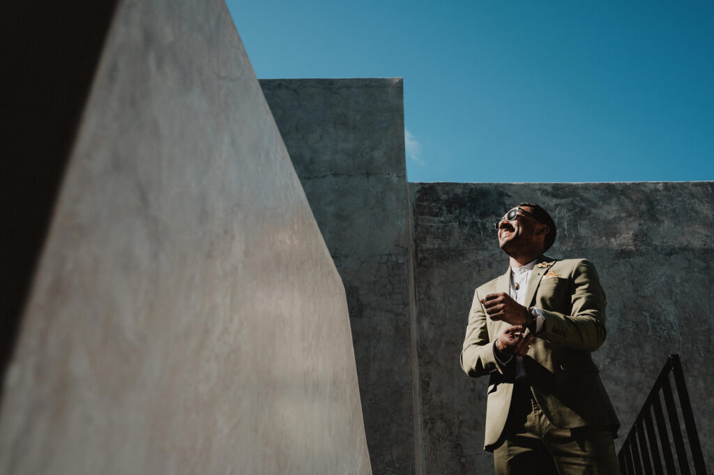 Groom adjusting cufflinks laughing outdoors at Frente al Punto Sayulita wedding, concrete walls blue sky