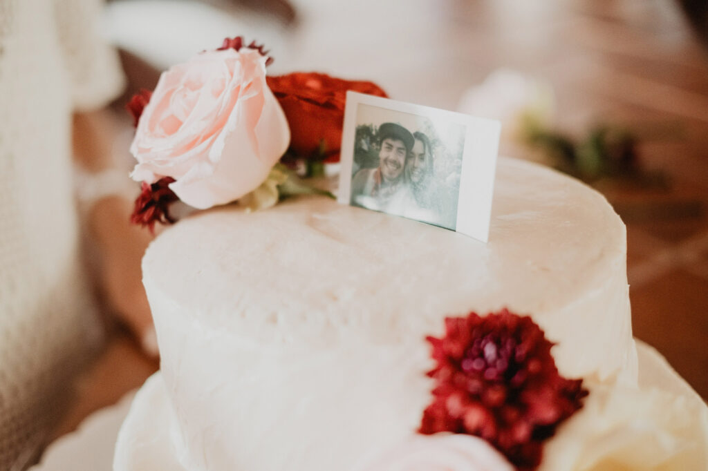 Two-tier white wedding cake with Polaroid photo topper and red flowers at Villa del Sol Sayulita reception