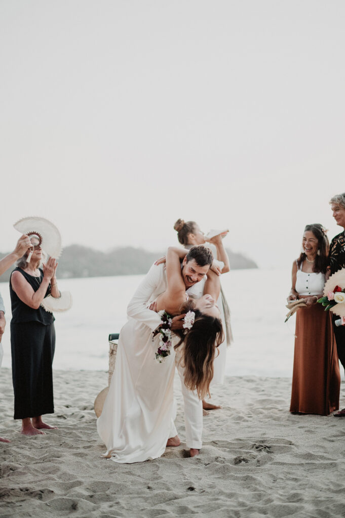Groom dips bride at beach wedding ceremony in Sayulita, Mexico, surrounded by laughing guests