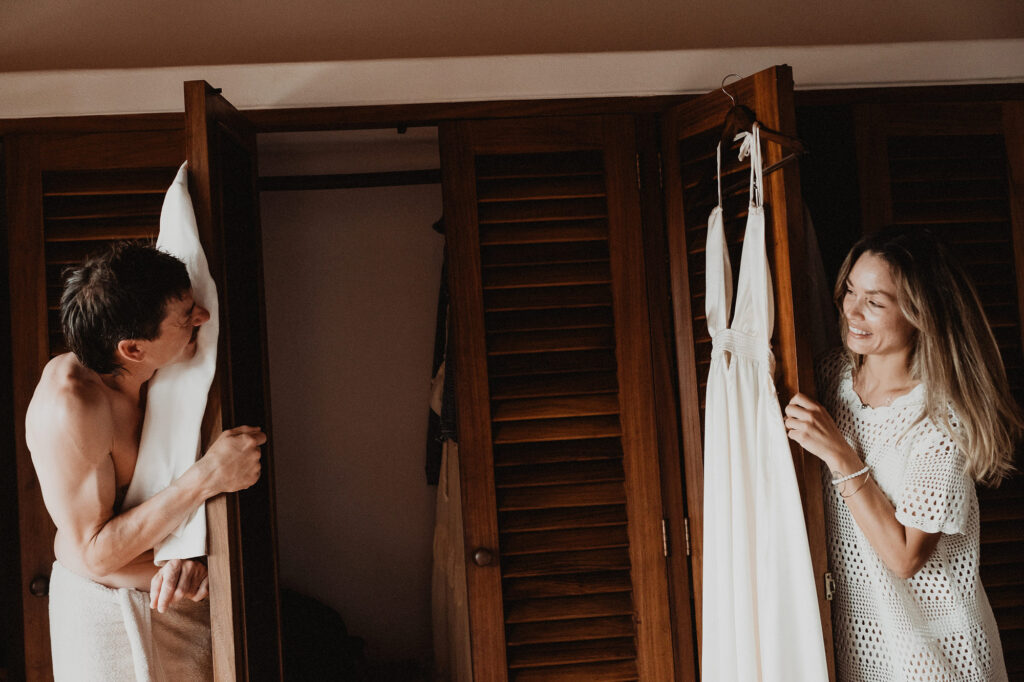Bride smiling at wedding dress on hanger while groom peeks from closet, Villa del Sol Sayulita wedding
