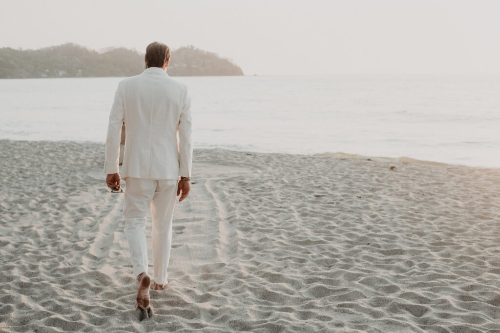 Groom in white linen suit walking barefoot on Sayulita beach at golden hour, Villa del Sol wedding