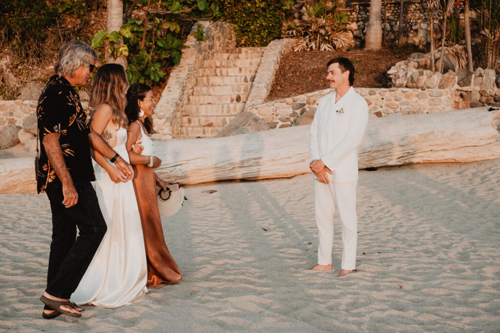 Groom in white suit laughing with bride and guests on sandy beach at Villa del Sol, Sayulita wedding ceremony