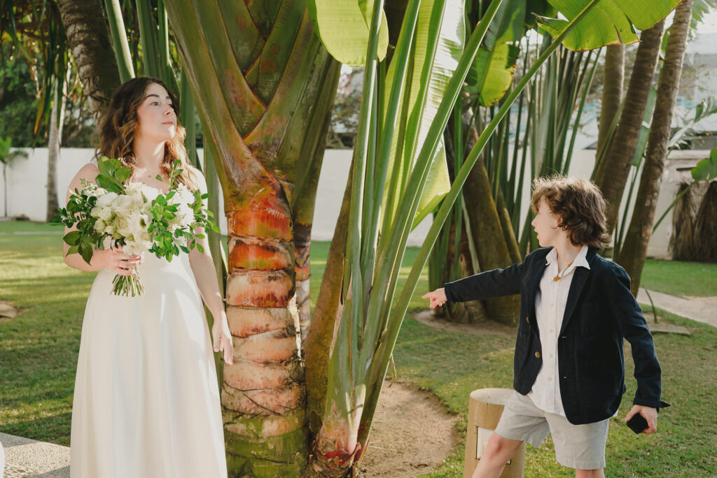 Bride holding white bouquet near tropical palms while a child points at her, wedding at Frente al Punto Sayulita