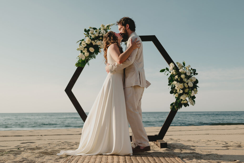 Bride and groom share first kiss at beach wedding ceremony, Sayulita Mexico, hexagonal floral arch backdrop