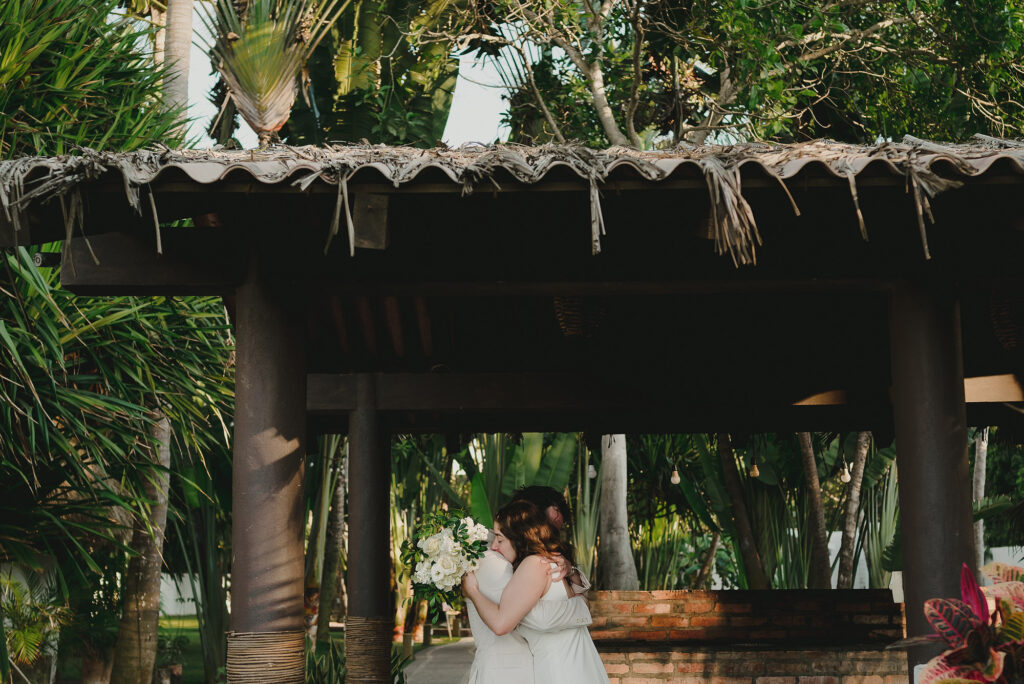 Two brides sharing first look embrace under palapa at Frente al Punto, Sayulita wedding
