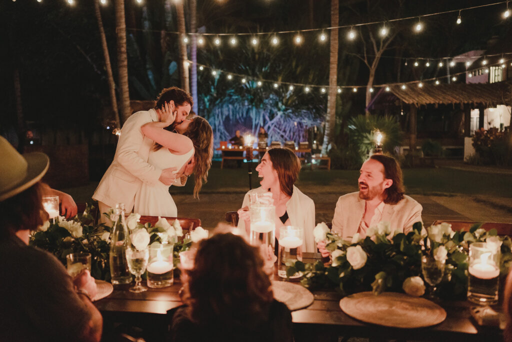 Couple kissing at candlelit wedding reception table in Sayulita, guests laughing, string lights overhead