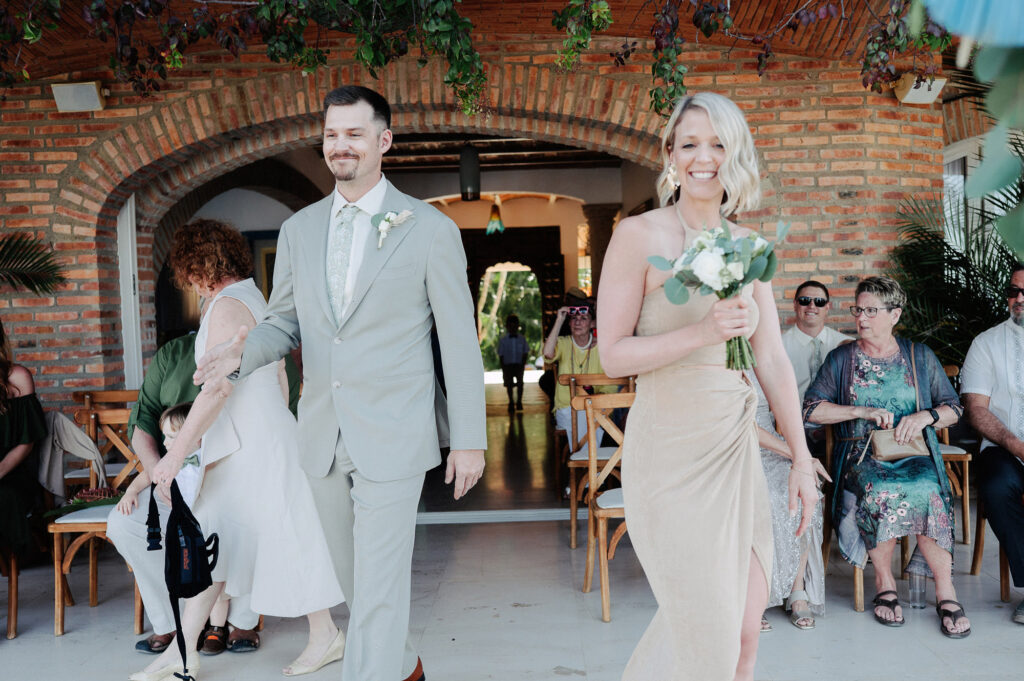 Groomsman and bridesmaid walk down aisle at Villa Amor Sayulita wedding ceremony, smiling with bouquet