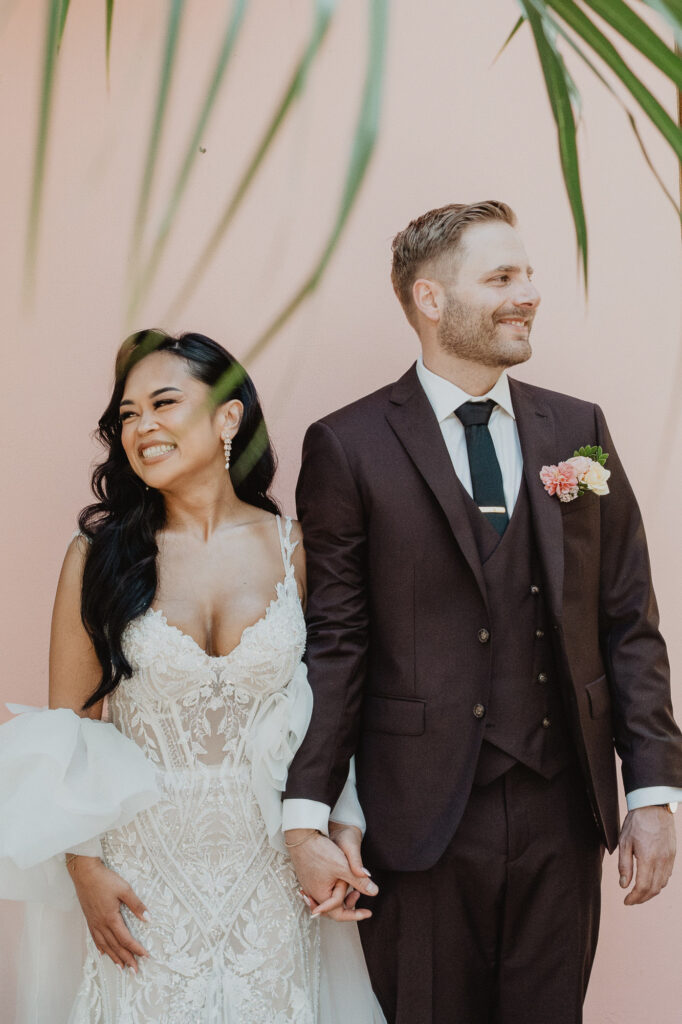 Bride and groom holding hands at Villa Valentin Sayulita wedding, lace gown and burgundy suit against pink wall