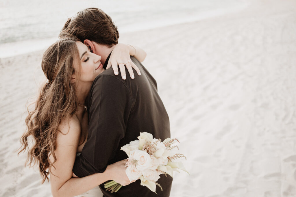 Bride holding bouquet embraces groom on sandy beach at St Regis Punta Mita elopement wedding